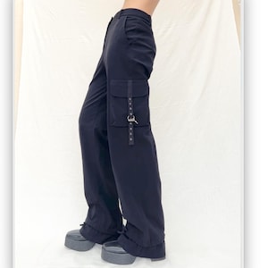 Puede incluir: Pantalones cargo negros de pierna ancha. Los pantalones tienen un bolsillo grande en el lateral con una correa y hebilla. Los pantalones se llevan con zapatos de plataforma.