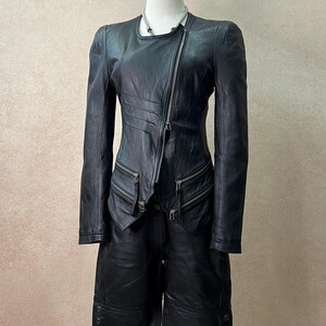 May include: A black leather jacket and matching leather shorts set. The jacket features an asymmetrical silver zipper, long sleeves, and two zippered pockets. The shorts are knee-length with three buttons on the outer hem of each leg opening.