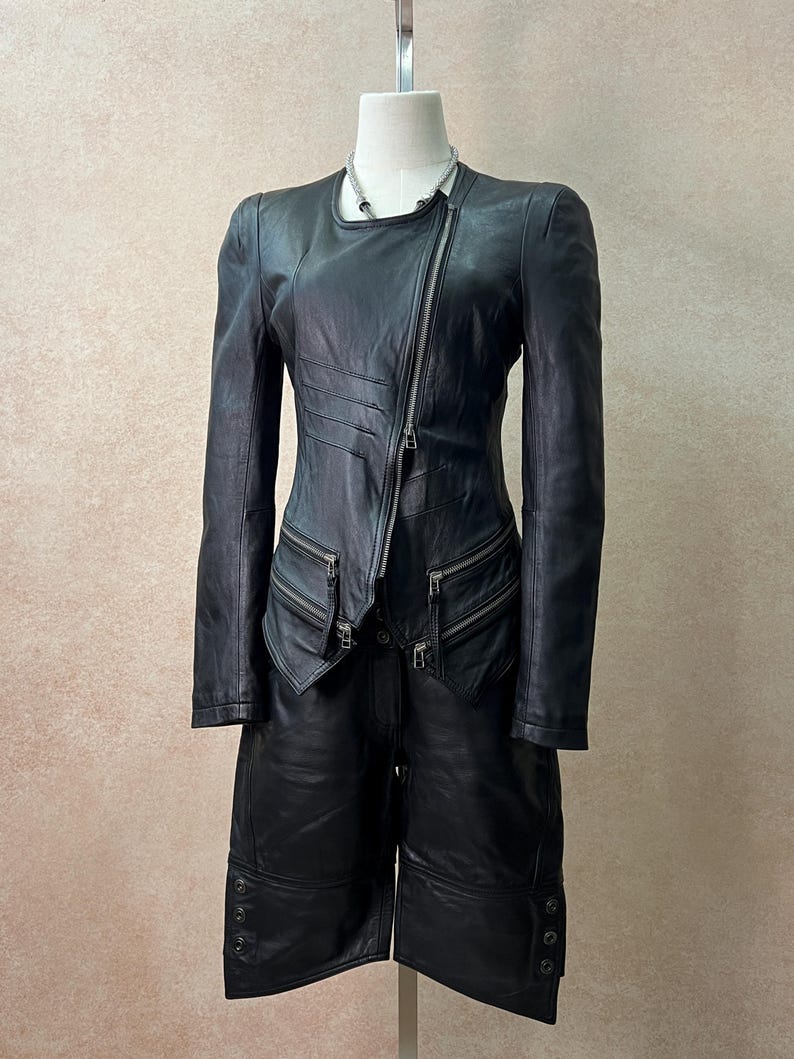May include: A black leather jacket and matching leather shorts set. The jacket features an asymmetrical silver zipper, long sleeves, and two zippered pockets. The shorts are knee-length with three buttons on the outer hem of each leg opening.