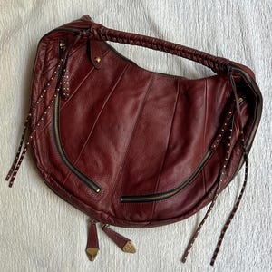 Peut inclure: Un sac hobo en cuir rouge foncé en forme de croissant avec une poignée tressée, plusieurs poches extérieures zippées dotées de longues tirettes en cuir cloutées et des détails en métal doré. Le sac présente une finition texturée et des panneaux verticaux.