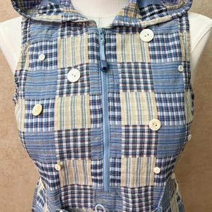 Puede incluir: Un chaleco sin mangas con capucha que presenta un diseño de patchwork con cuadrados de tela a cuadros en azul, blanco y beige. La prenda incluye una cremallera central azul claro, un cordón en la cintura y varios botones blancos.