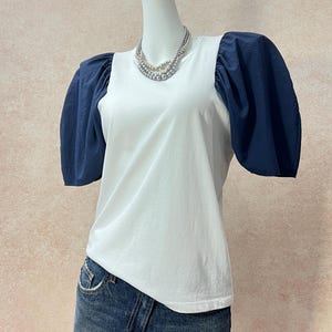 May include: A white t-shirt featuring voluminous navy blue puff sleeves and a crew neckline. The shirt is styled with a multi-strand silver-toned beaded necklace and a blue denim skirt. The garment has a solid white front panel and dark sleeves.