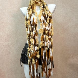 May include: A long, textured scarf featuring a mix of white, tan, and dark brown fuzzy yarn strands. The scarf has an open, knotted construction with long, dangling fringe elements that hang down from the main body of the piece.