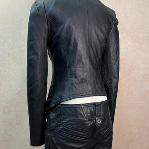 May include: A black leather jacket with a high collar and long sleeves, paired with matching black leather pants. The jacket features a fitted silhouette with visible seam detailing, while the pants include a metal button and pocket accents.
