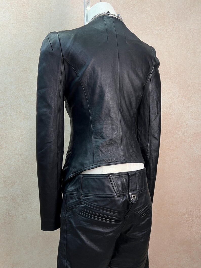 May include: A black leather jacket with a high collar and long sleeves, paired with matching black leather pants. The jacket features a fitted silhouette with visible seam detailing, while the pants include a metal button and pocket accents.