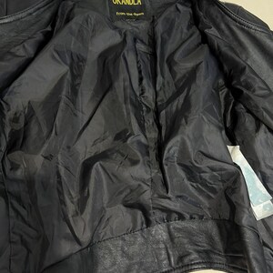 May include: A black leather jacket with a black interior lining. The inner neck label features the text OKANDLA and the phrase From the dream in yellow lettering. A white care tag is visible on the inner right side of the jacket interior.