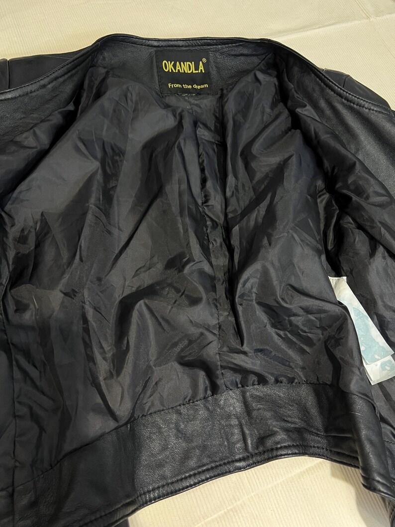 May include: A black leather jacket with a black interior lining. The inner neck label features the text OKANDLA and the phrase From the dream in yellow lettering. A white care tag is visible on the inner right side of the jacket interior.