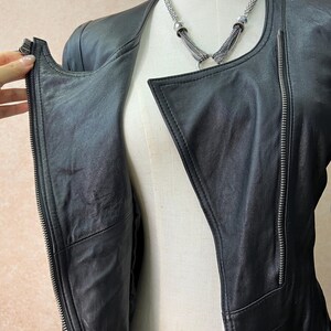 May include: A black leather jacket featuring an asymmetrical front zipper closure, long sleeves, and a collarless neckline. The jacket is displayed on a mannequin and is paired with a silver-toned multi-strand chain necklace with metallic accents.
