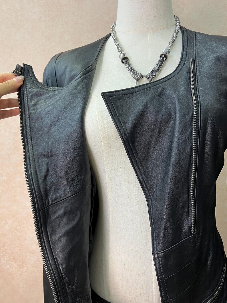 May include: A black leather jacket featuring an asymmetrical front zipper closure, long sleeves, and a collarless neckline. The jacket is displayed on a mannequin and is paired with a silver-toned multi-strand chain necklace with metallic accents.