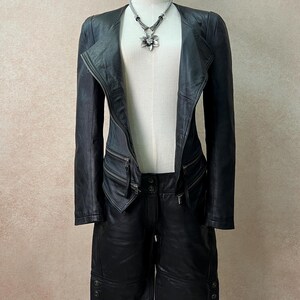 May include: A black leather jacket with an asymmetrical front zipper, long sleeves, and two zippered pockets at the waist, paired with matching black leather shorts featuring button details at the hem and a silver flower pendant necklace.