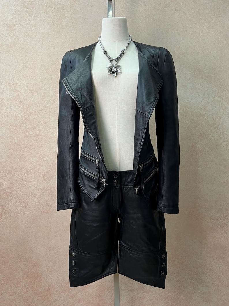 May include: A black leather jacket with an asymmetrical front zipper, long sleeves, and two zippered pockets at the waist, paired with matching black leather shorts featuring button details at the hem and a silver flower pendant necklace.