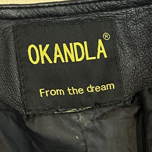 May include: A black leather garment featuring a rectangular black fabric label stitched onto the interior. The label displays the yellow text OKANDLA with a registered trademark symbol and the phrase From the dream in yellow lettering below.