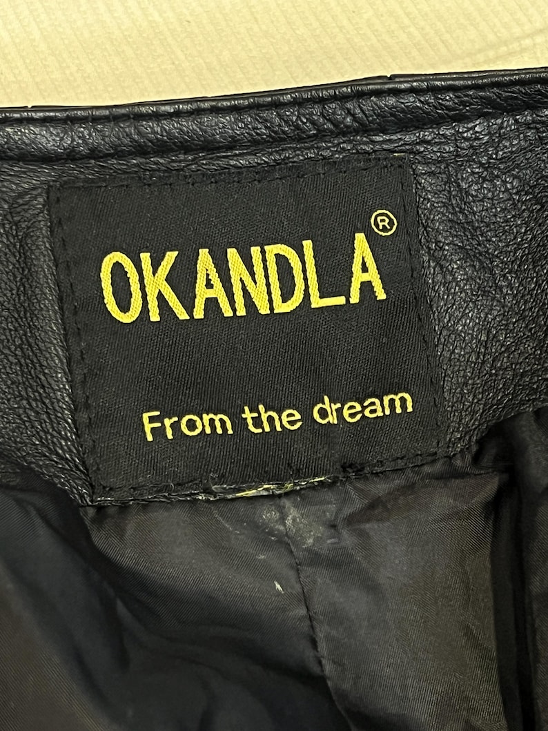 May include: A black leather garment featuring a rectangular black fabric label stitched onto the interior. The label displays the yellow text OKANDLA with a registered trademark symbol and the phrase From the dream in yellow lettering below.