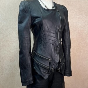 May include: A black leather jacket featuring a diagonal front zipper closure, long sleeves, and a round neckline. The jacket includes horizontal stitched detailing on the front torso and two zippered pockets positioned at the lower waist area.