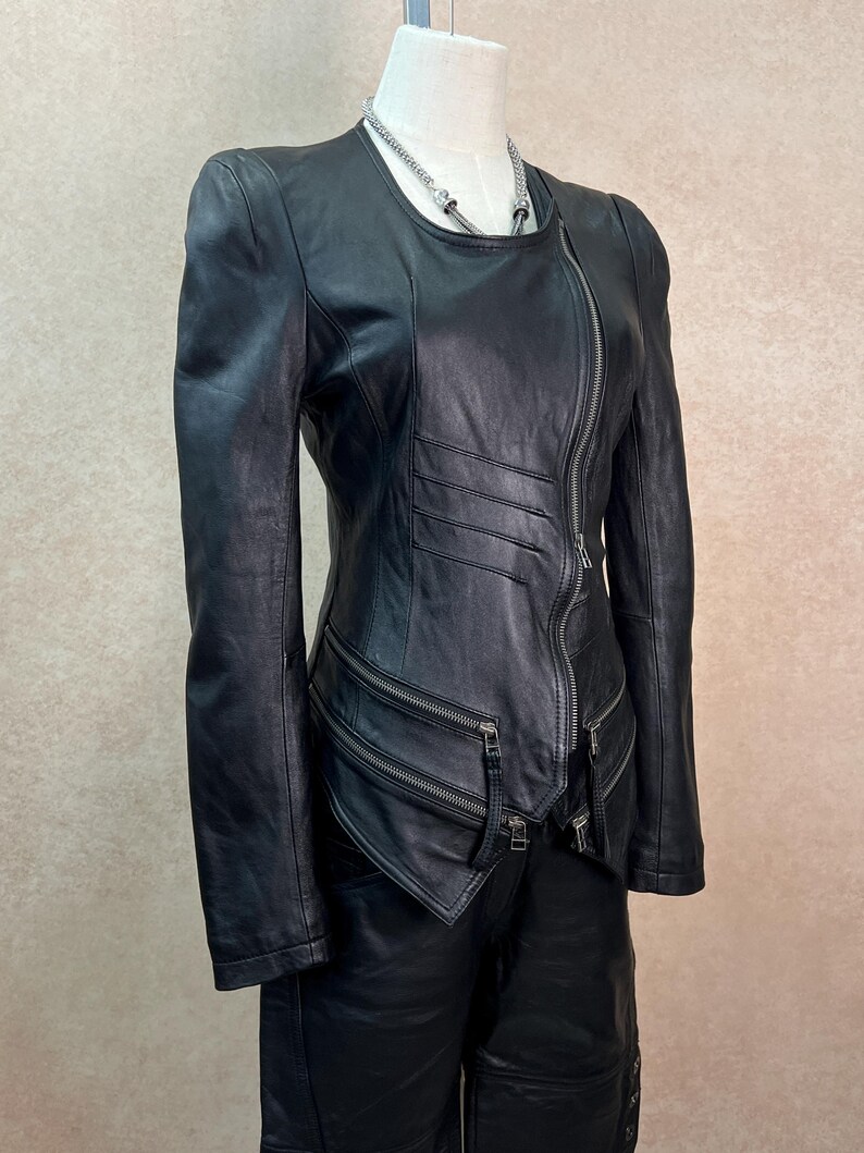 May include: A black leather jacket featuring a diagonal front zipper closure, long sleeves, and a round neckline. The jacket includes horizontal stitched detailing on the front torso and two zippered pockets positioned at the lower waist area.