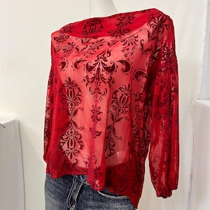 May include: A long-sleeved red blouse featuring a sheer, lightweight fabric with a dark red velvet damask pattern. The top has a wide neckline and gathered cuffs at the wrists, paired with a dark blue denim skirt with a visible front pocket.