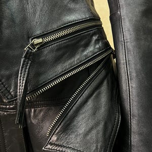 May include: A black leather jacket featuring multiple silver-toned metal zippers. The garment includes a horizontal zippered pocket and an angled zippered pocket with a leather pull tab attached to the slider, showcasing detailed stitching.
