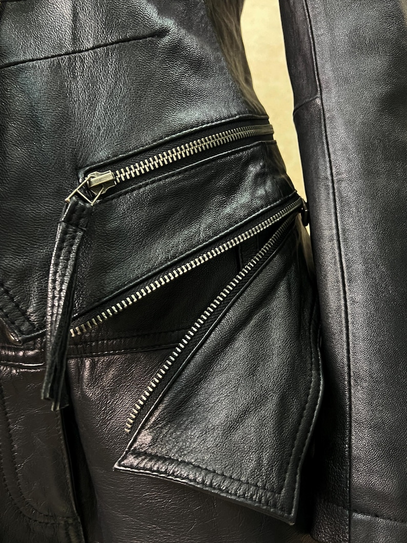 May include: A black leather jacket featuring multiple silver-toned metal zippers. The garment includes a horizontal zippered pocket and an angled zippered pocket with a leather pull tab attached to the slider, showcasing detailed stitching.