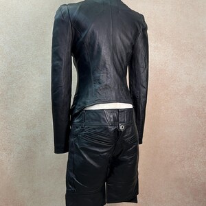 May include: A black leather jacket and matching black leather shorts set. The jacket features long sleeves and a fitted silhouette. The shorts have a mid-rise waist with a single silver-toned button closure, visible back pockets, and an asymmetrical hemline.