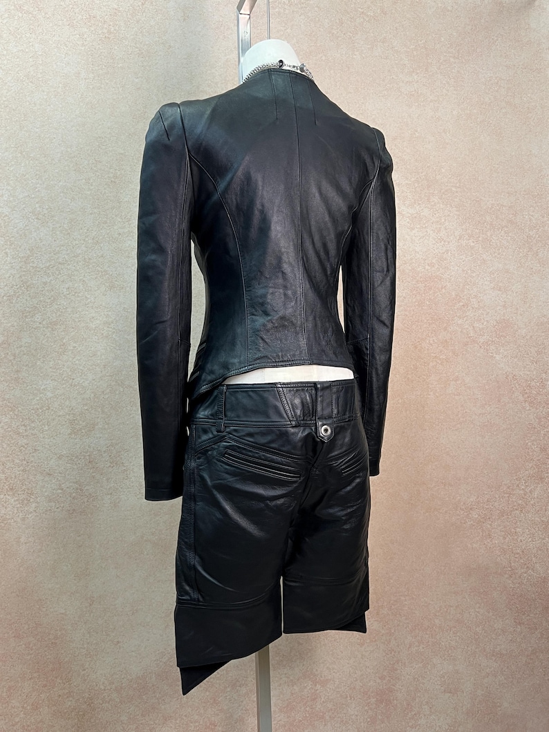 May include: A black leather jacket and matching black leather shorts set. The jacket features long sleeves and a fitted silhouette. The shorts have a mid-rise waist with a single silver-toned button closure, visible back pockets, and an asymmetrical hemline.