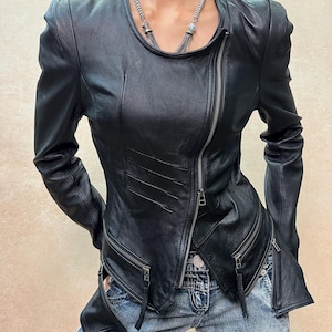 May include: A black leather jacket featuring a round neckline, long sleeves, and an asymmetrical front zipper closure. The jacket includes two zippered pockets at the waist, decorative diagonal stitching on the front panel, and a silver-toned chain necklace.