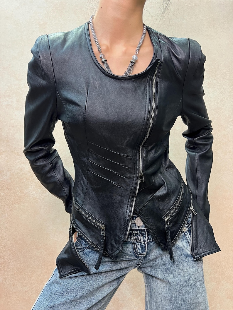 May include: A black leather jacket featuring a round neckline, long sleeves, and an asymmetrical front zipper closure. The jacket includes two zippered pockets at the waist, decorative diagonal stitching on the front panel, and a silver-toned chain necklace.