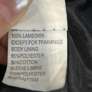 May include: A white garment care label with black text reading: OUTER SHELL 100% LAMBSKIN EXCEPT FOR TRIMMINGS, BODY LINING 65% POLYESTER 35% COTTON, SLEEVE LINING 100% POLYESTER, and RN(USA) 77302, attached to a black fabric interior lining.