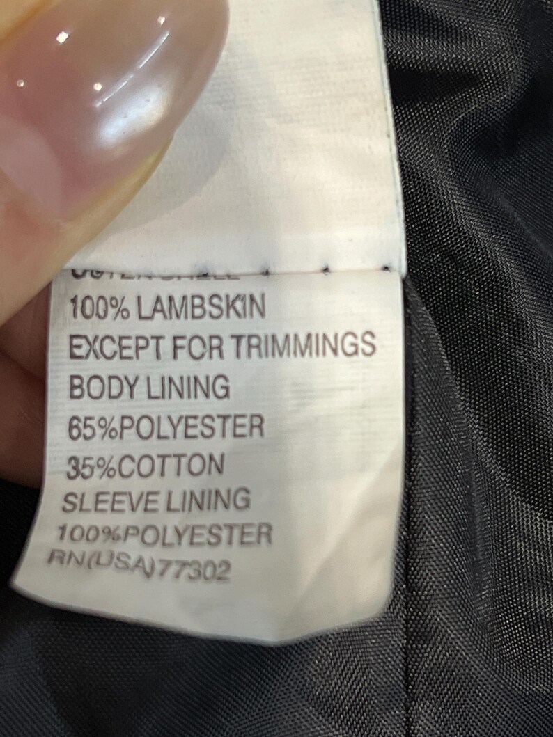 May include: A white garment care label with black text reading: OUTER SHELL 100% LAMBSKIN EXCEPT FOR TRIMMINGS, BODY LINING 65% POLYESTER 35% COTTON, SLEEVE LINING 100% POLYESTER, and RN(USA) 77302, attached to a black fabric interior lining.