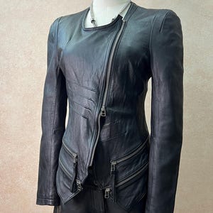 May include: A black leather jacket featuring an asymmetrical silver zipper closure, a round neckline, and long sleeves. The jacket includes horizontal stitched detailing on the right side and two zippered pockets at the waist with silver hardware.