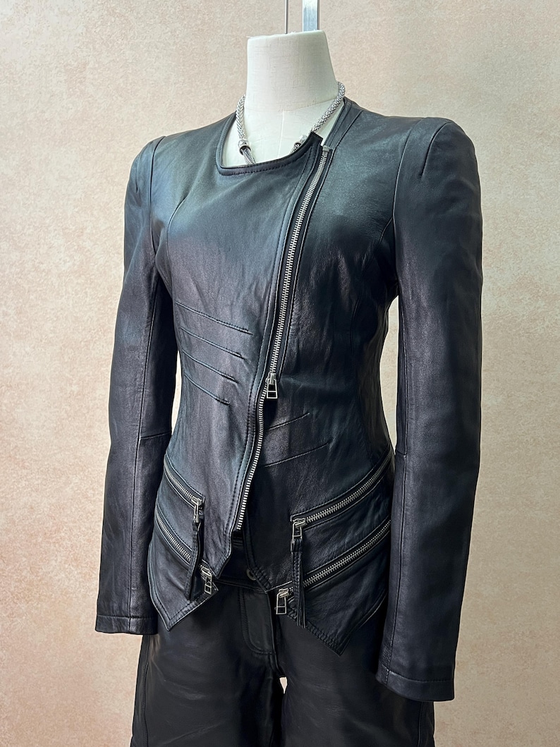 May include: A black leather jacket featuring an asymmetrical silver zipper closure, a round neckline, and long sleeves. The jacket includes horizontal stitched detailing on the right side and two zippered pockets at the waist with silver hardware.