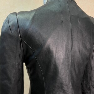 May include: A black leather jacket featuring a smooth finish, structured seams along the back, and long sleeves. The jacket has a high neckline and is displayed on a mannequin, with a silver-toned chain necklace visible around the neck area.