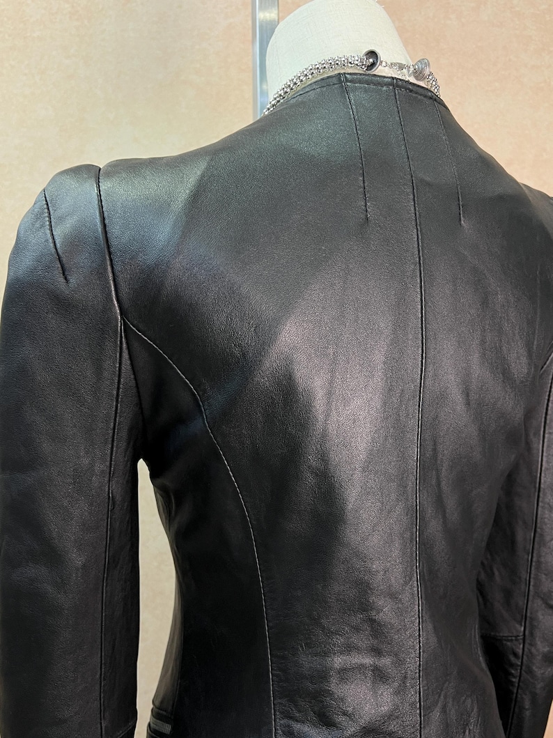 May include: A black leather jacket featuring a smooth finish, structured seams along the back, and long sleeves. The jacket has a high neckline and is displayed on a mannequin, with a silver-toned chain necklace visible around the neck area.