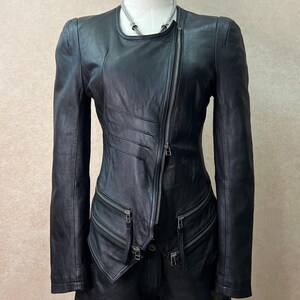 May include: A black leather jacket featuring an asymmetrical front zipper closure, long sleeves, and multiple zippered pockets at the waist. The jacket has a round neckline and decorative horizontal stitched pleats on the right side of the chest.