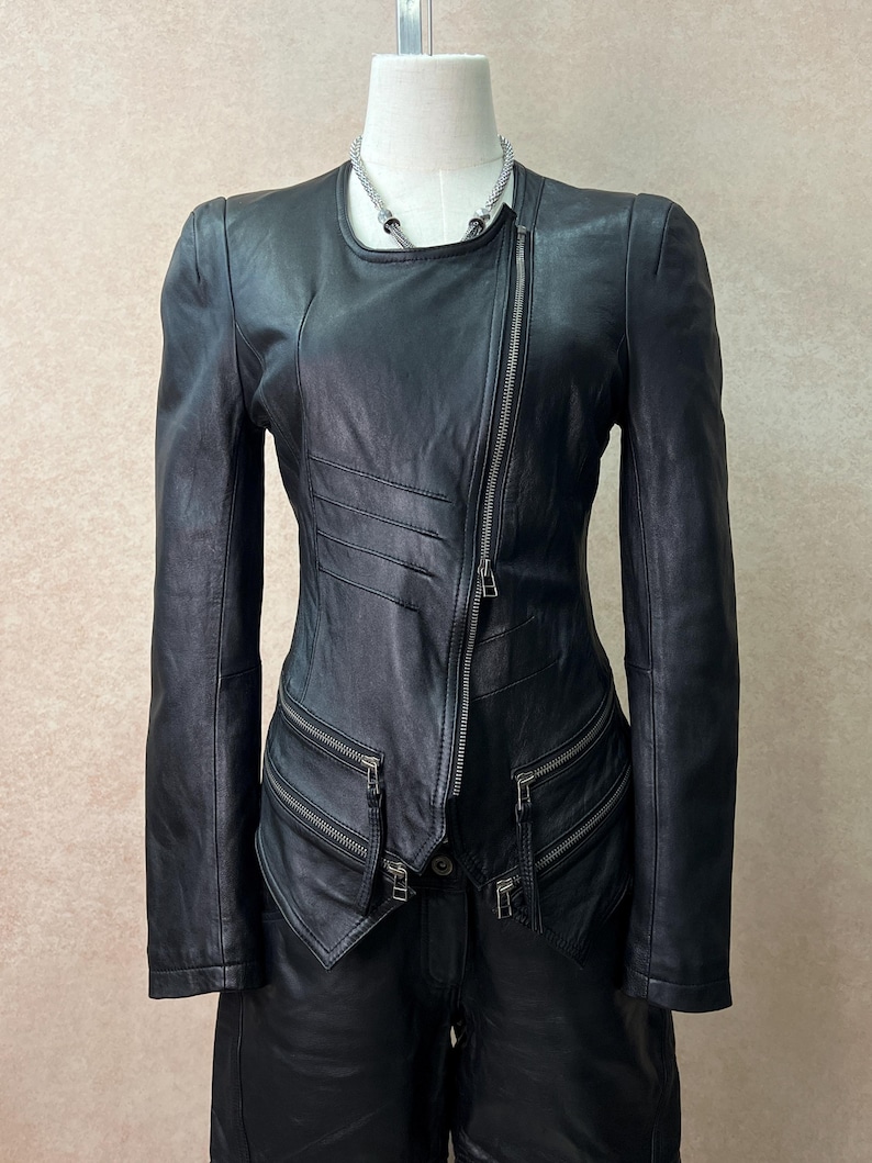 May include: A black leather jacket featuring an asymmetrical front zipper closure, long sleeves, and multiple zippered pockets at the waist. The jacket has a round neckline and decorative horizontal stitched pleats on the right side of the chest.