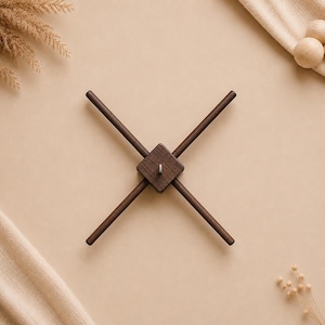 Può includere: Una gruccia per giostrina in legno marrone scuro con un blocco centrale quadrato, quattro bracci radianti e un piccolo gancio metallico che sporge dal centro. La struttura è simmetrica e forma una X con aste di legno dritte e lisce.