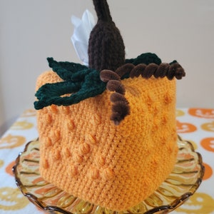 May include: A crocheted pumpkin-shaped tissue box cover with a brown stem, green leaves, and a brown pipe cleaner accent. The cover is sitting on a glass cake stand.