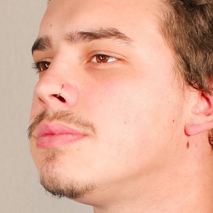 May include: A close-up of a person's face with a nose ring. The person has short brown hair and is looking to the side.