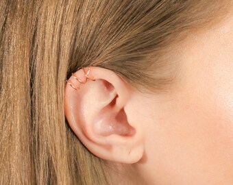 May include: A close-up of a woman's ear with three small, gold-colored ear cuffs. The cuffs are made of thin wire and are worn on the upper part of the ear.