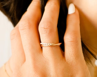 May include: A close-up of a hand wearing a delicate gold ring with a twisted design. The ring is made of two bands, one with a textured surface and the other with a smooth surface. The ring is worn on the ring finger of the left hand.