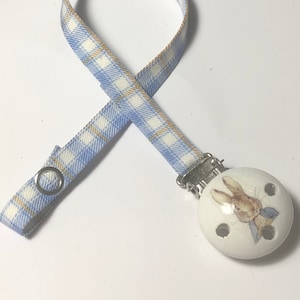 Peut inclure: Un attache-sucette en tissu à carreaux bleus et blancs avec un charme de sucette en céramique blanche représentant une illustration de Lapin de Pâques.