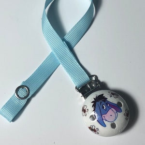 Könnte beinhalten: Ein hellblauer Schnullerclip mit einem runden weißen Holzanhänger mit einem Cartoon-Bild von Eeyore aus Winnie Puuh. Der Anhänger hat braune Bienendesigns am Rand.