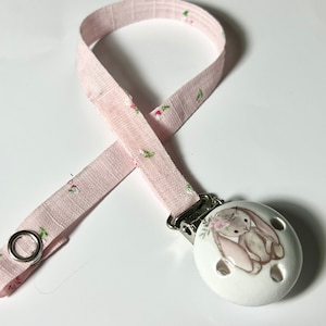 May include: A pink and white pacifier clip with a floral pattern and a bunny design. The clip is made of fabric and has a metal ring.