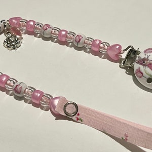 Peut inclure: Attache-sucette en perles rose et blanche avec un clip en céramique blanche représentant une souris et le nom "FREYA" écrit en lettres noires.