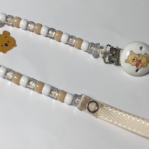 Peut inclure: Un attache-sucette en perles blanches et beiges avec un charme Winnie l'ourson et un attache-sucette en céramique blanche Winnie l'ourson. L'attache est fixée à un ruban beige.