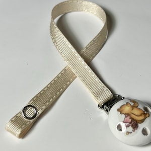 Peut inclure: Un ruban de satin crème attaché à un clip de sucette en céramique blanche avec un motif Winnie l'ourson et Porcinet.