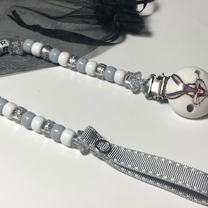 May include: A baby pacifier with a white, gray, and silver beaded chain. The pacifier has a white nipple with a Bugs Bunny graphic. The chain spells "CASPER" with white letter beads. A gray ribbon loop is attached.