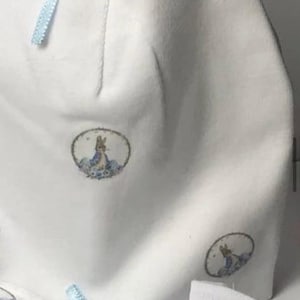 Może przedstawiać: Biały zestaw dla niemowląt, składający się z czapeczki i pasujących akcesoriów, ozdobiony okrągłą ilustracją królika w niebiesko-zielonym otoczeniu kwiatowym. Jasnoniebieskie wstążki dodają zestawowi delikatności.