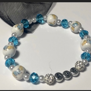 May include: A white beaded bracelet with blue crystal beads and white beads with a cartoon bear design. The bracelet has the name "AVA" spelled out in silver beads.