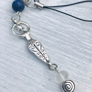 May include: A silver charm featuring a stylized female figure with decorative patterns, a blue bead, and a spiral charm. The charm is attached to a black cord, suitable for use as a keychain or accessory.