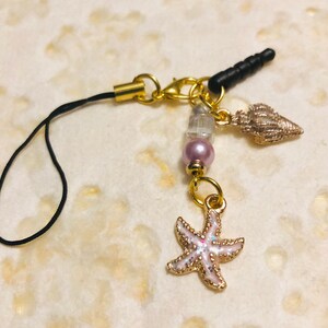 May include: A gold charm with a black phone strap, a pink pearl, a clear crystal, a gold seashell, and a gold starfish. The charm is perfect for adding a touch of whimsy to your phone or bag.